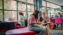 Two young women sitting on a red chair chatting and holding cheat sheets in their hands