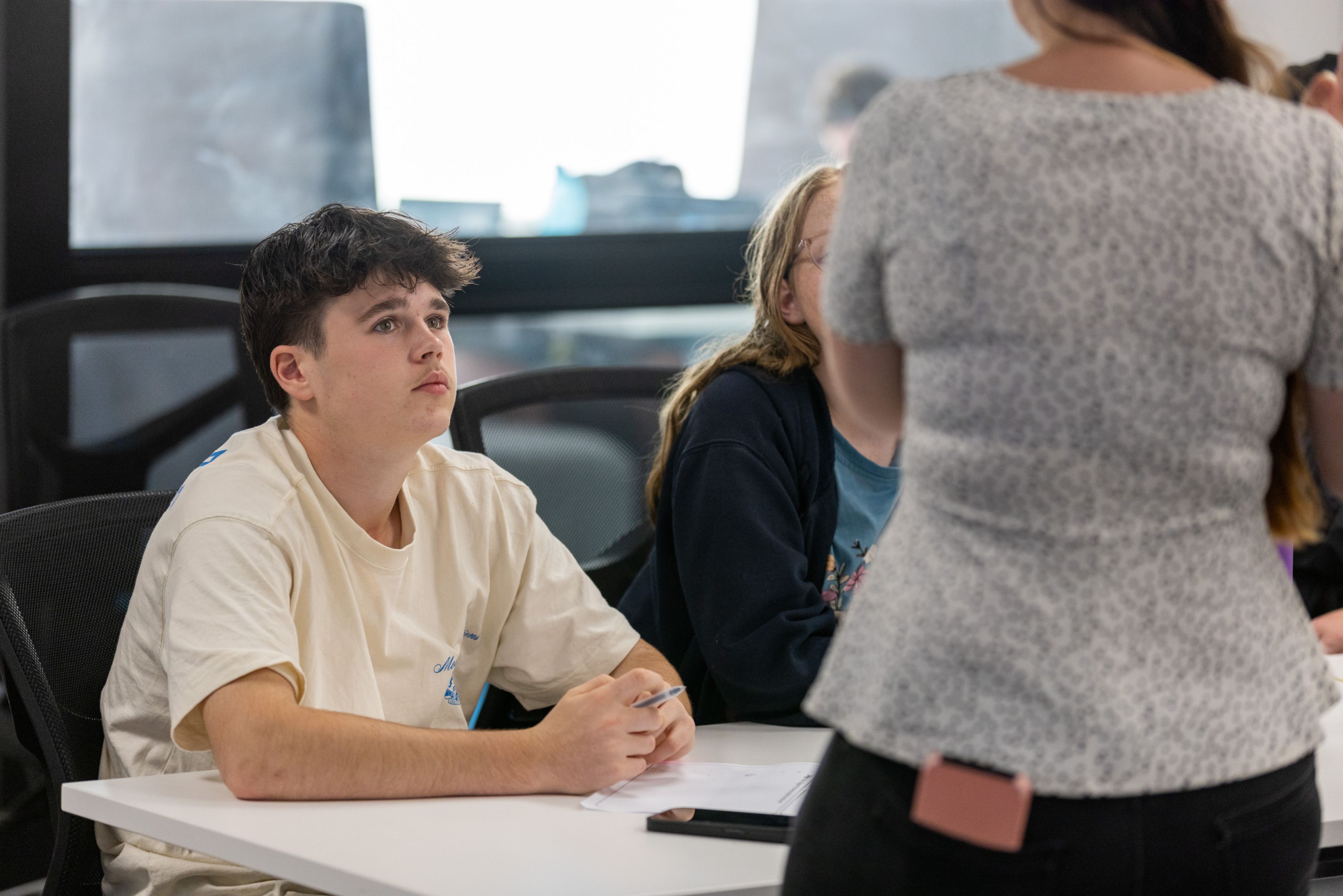 A student sitting at a classroom desk, listening to a teacher during a workshop.