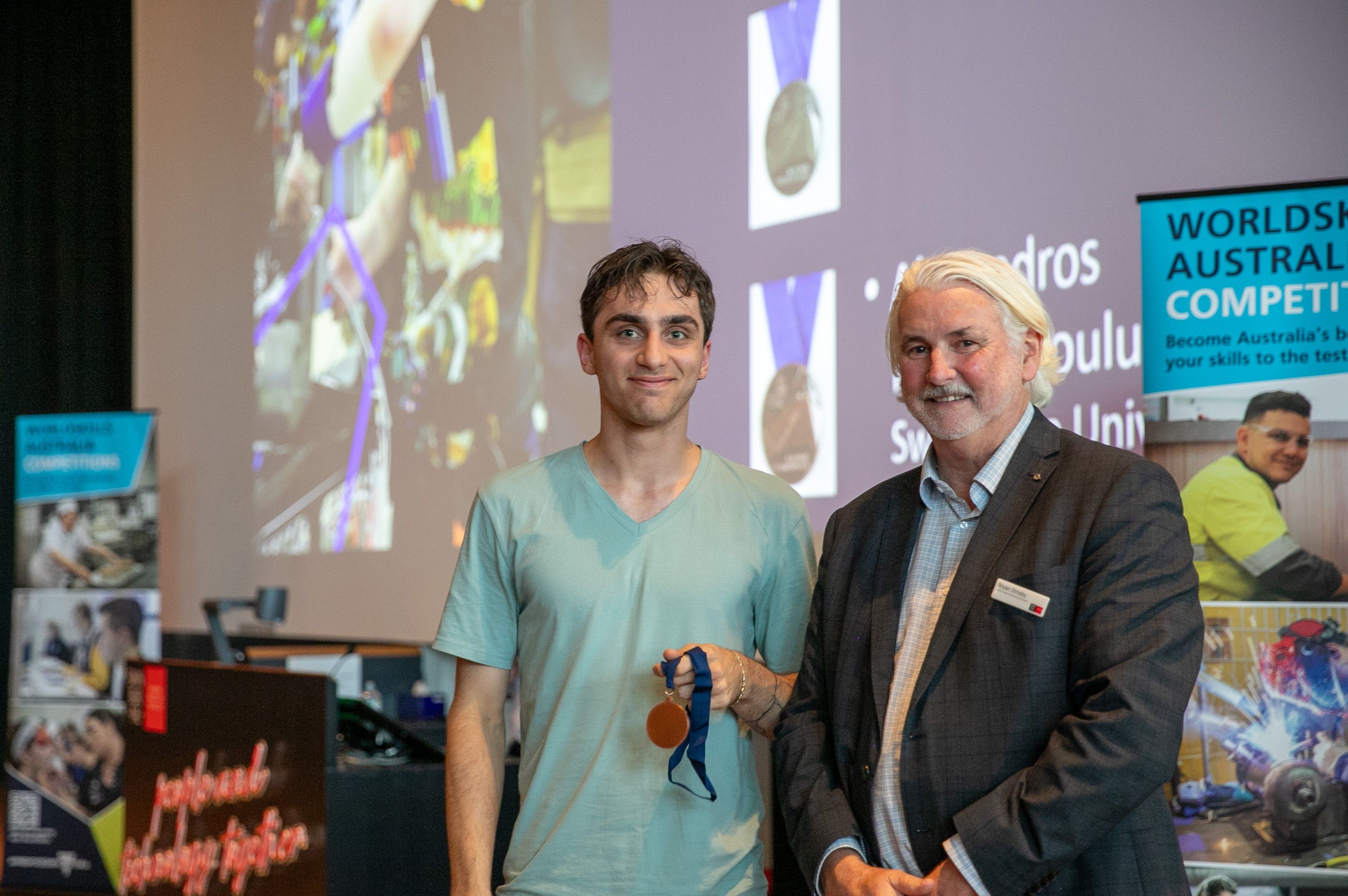 Male student standing next to teacher, holding a bronze medal and smiling. 