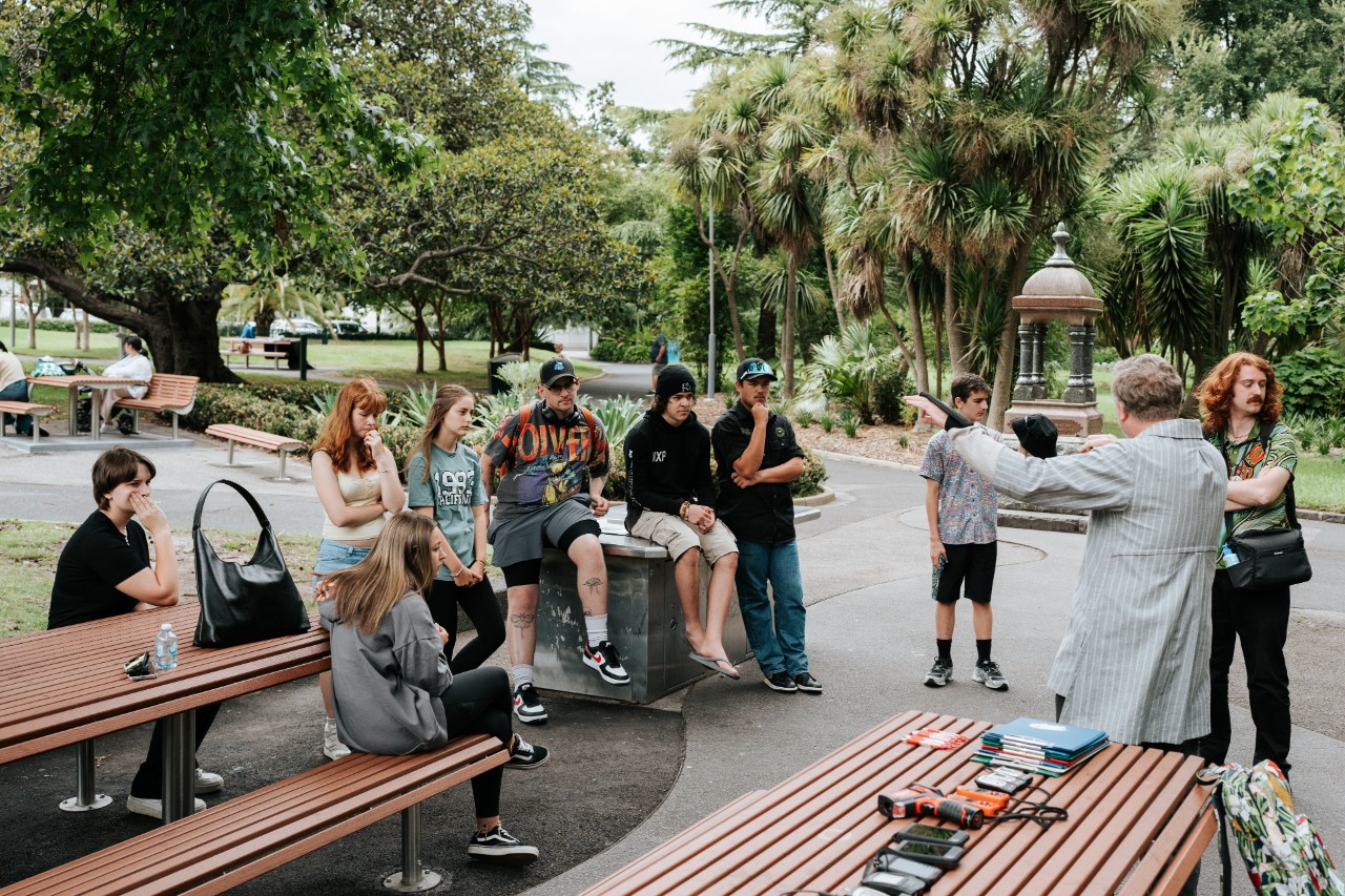 Young adult students sitting on park benches and BBQs in a park, listening to an older man speak.