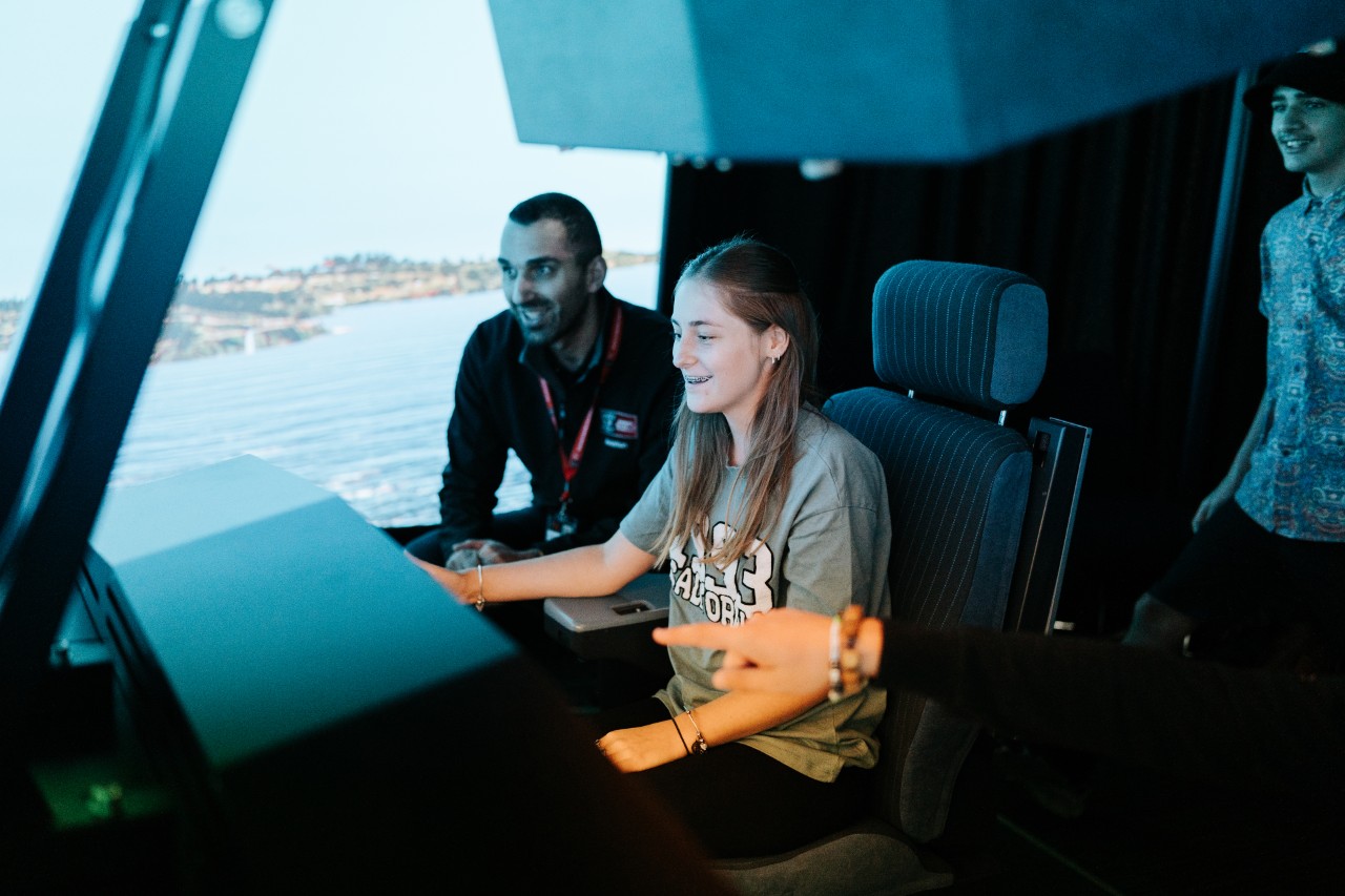 Female young adult sitting in a chair in front of a pilot monitor, with male teacher on her right.