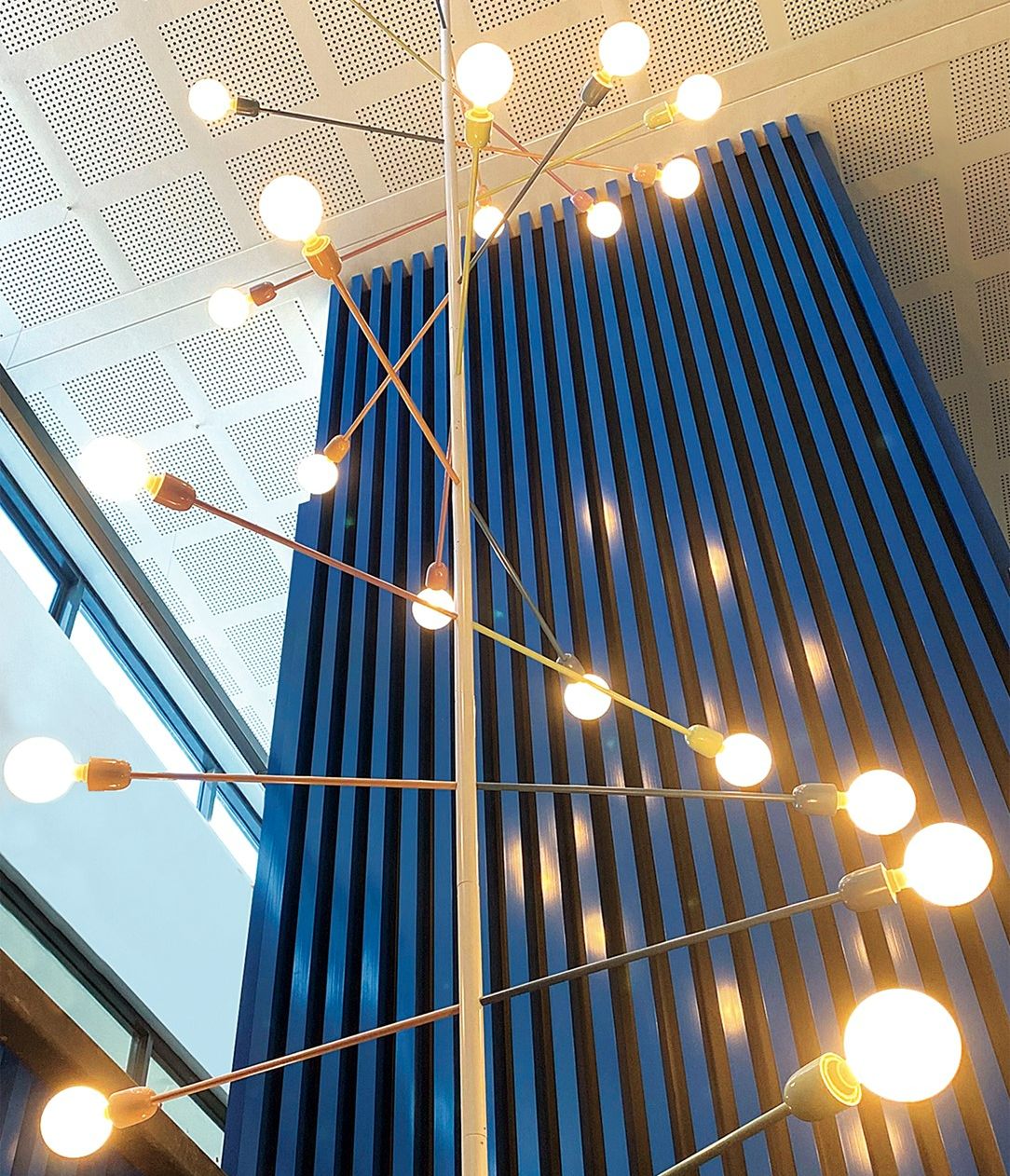 A DNA spiral shaped light feature hanging from a tall ceiling. Metal rods with warmly lit light globes on each end represent DNA’s base pairs, giving the light its double helix appearance.