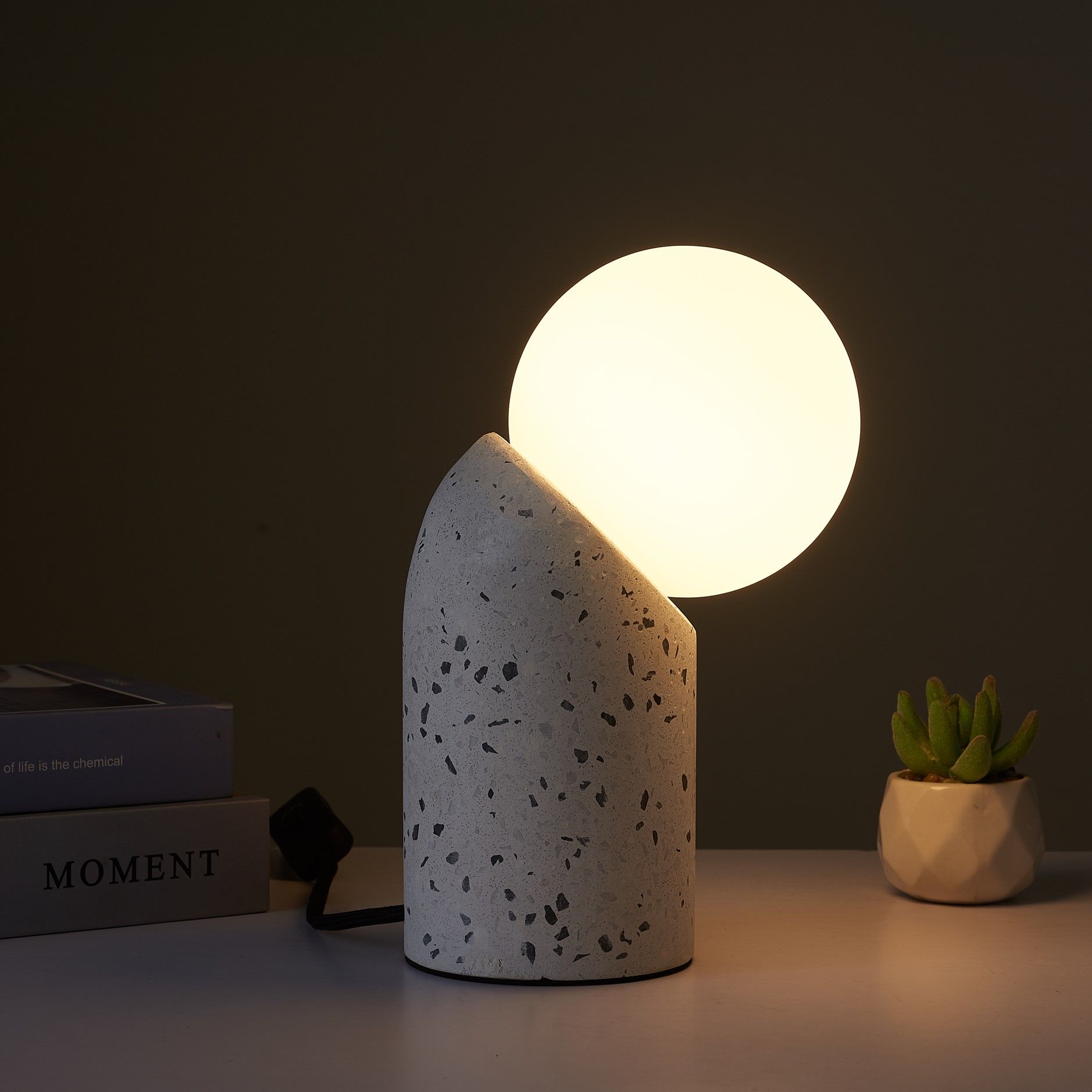 A lamp in a dark room warmly lighting a desk. The lamp is made up of a white, speckled cylindrical base, curving at the top to create an angled surface for the attached spherical light shade. 