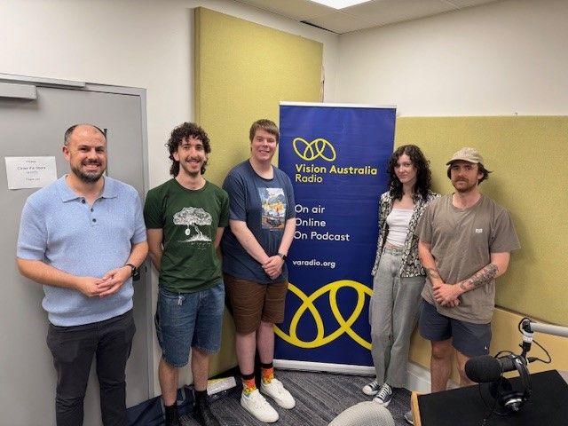 Five people smiling and standing in front of a blue and yellow Vision Australia Radio banner.