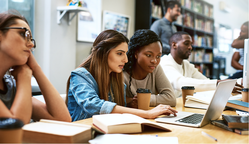 two students work at laptop in a library setting. 