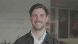 Photograph of Nathan Loriente from Metro Trains. Nathan wears a white collared shirt and jacket and is standing in front of an office.