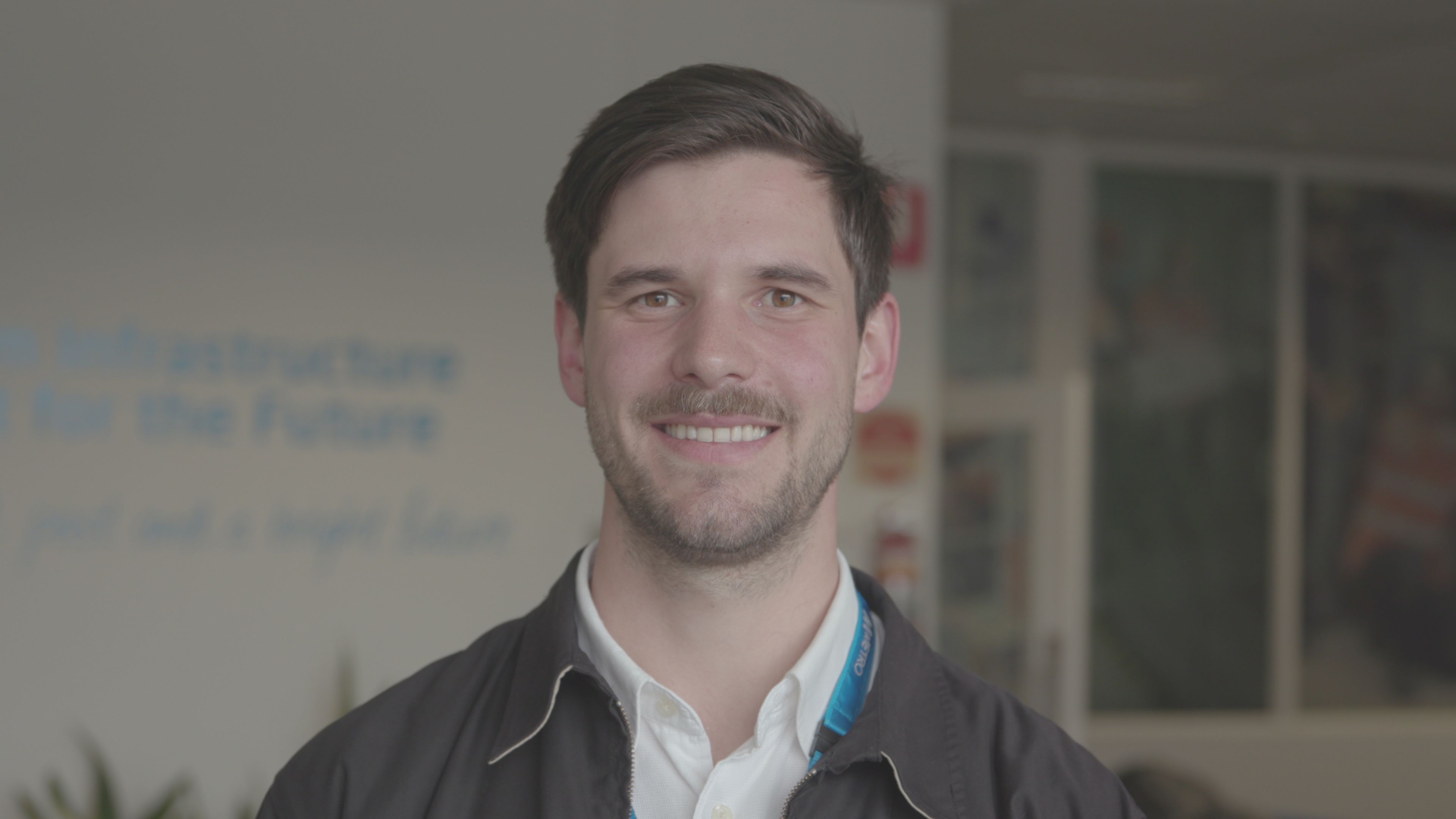 Photograph of Nathan Loriente from Metro Trains. Nathan wears a white collared shirt and jacket and is standing in front of an office.