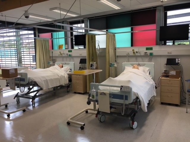 Two full-body patient simulators lay in two hospital beds side by side. There is hospital equipment around the beds.