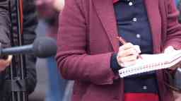 Female journalist at news conference or media event, writing notes, holding a microphone.