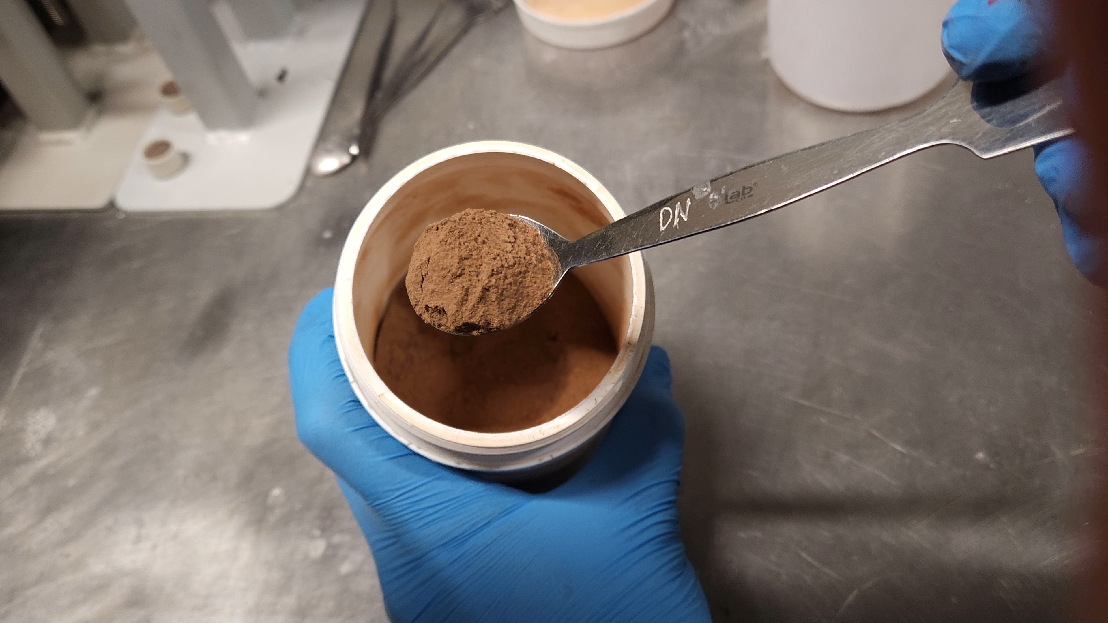 A spoonful of fine, red dust is held above a container holding the material. The dust is regolith simulant, made to mimic the surface composition found at Gale Crater on Mars.