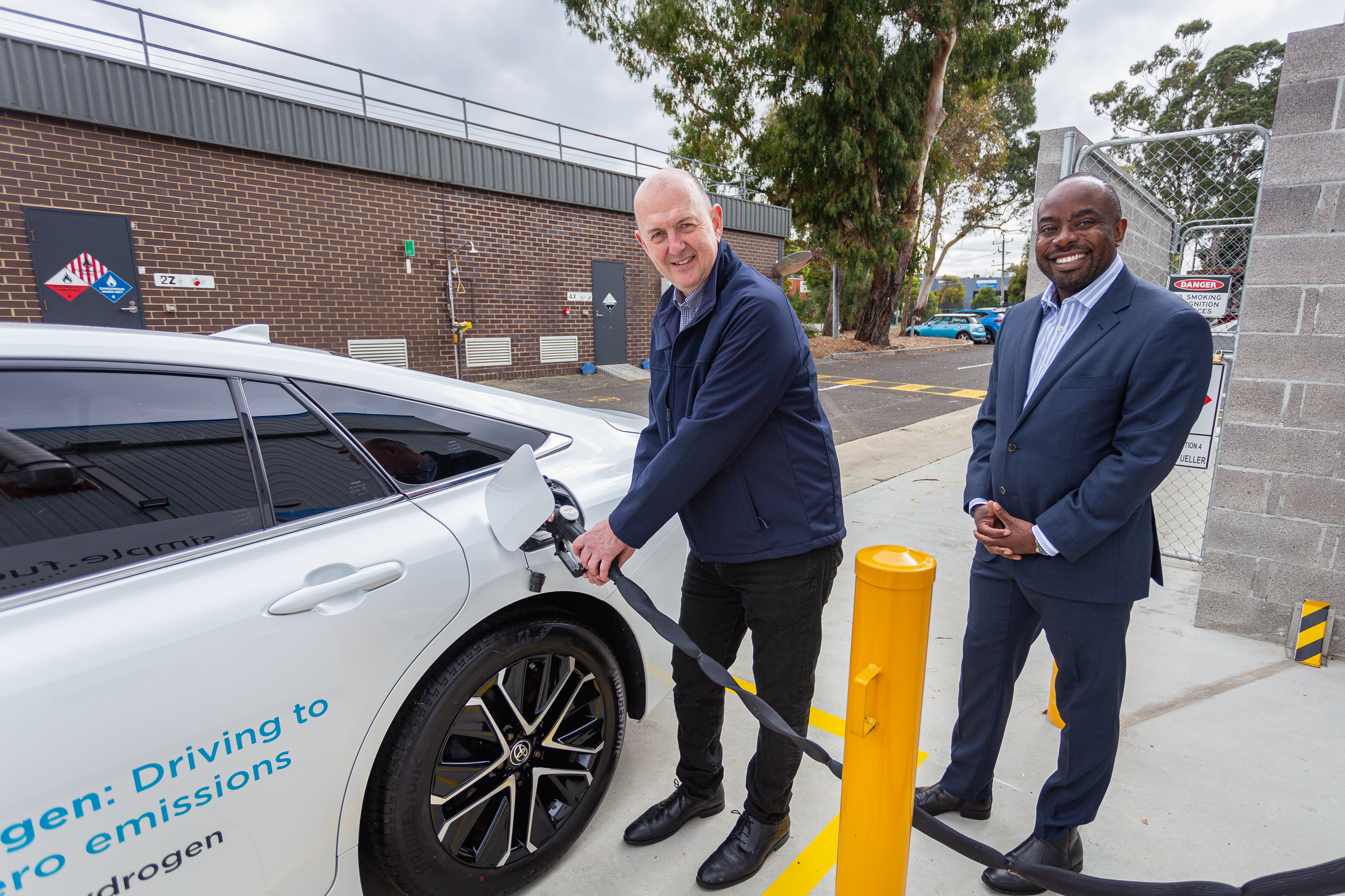 Two men smiling for the camera, man on left holding a petrol pump, putting petrol into a white car, man on right in a suit holding his hands smiling at the camera.