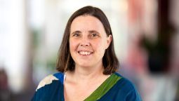 Woman with short brown hair in a blue, green and whit blouse, facing front on, smiling at the camera 