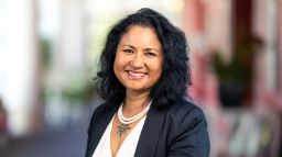 Woman with white top and back cardigan, black hair, nose ring and tattoo on her chest smiling at the camera.