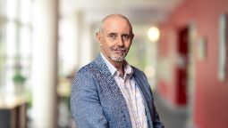 headshot of man with white striped shirt and grey blazer looking at camera