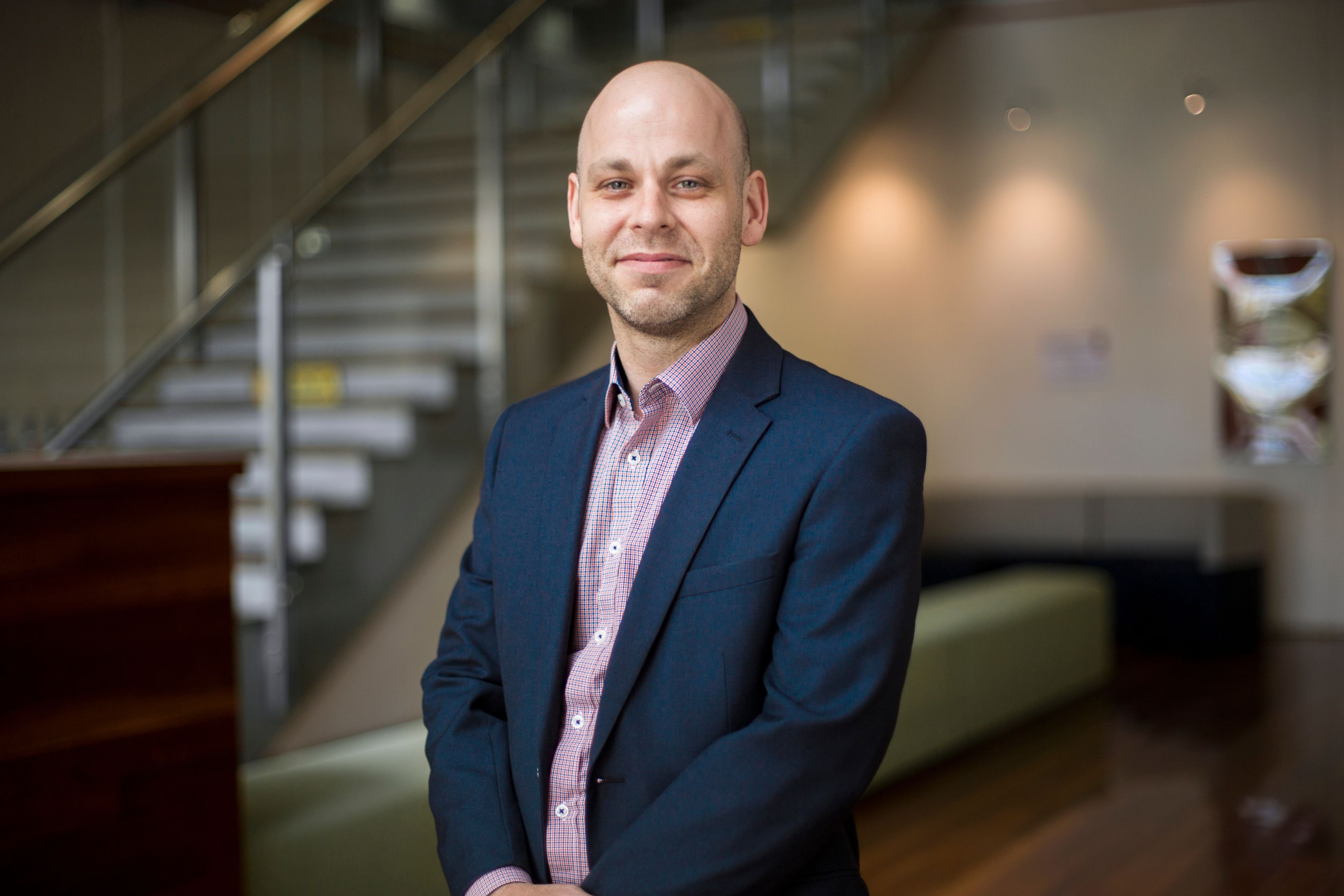 Travis Keating wearing suit attire poses with hands clasped in front in the foyer of the SPS building
