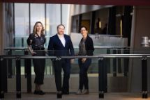 Headshot of two female professionals and one male professional on a bridge against a glass wall background. 