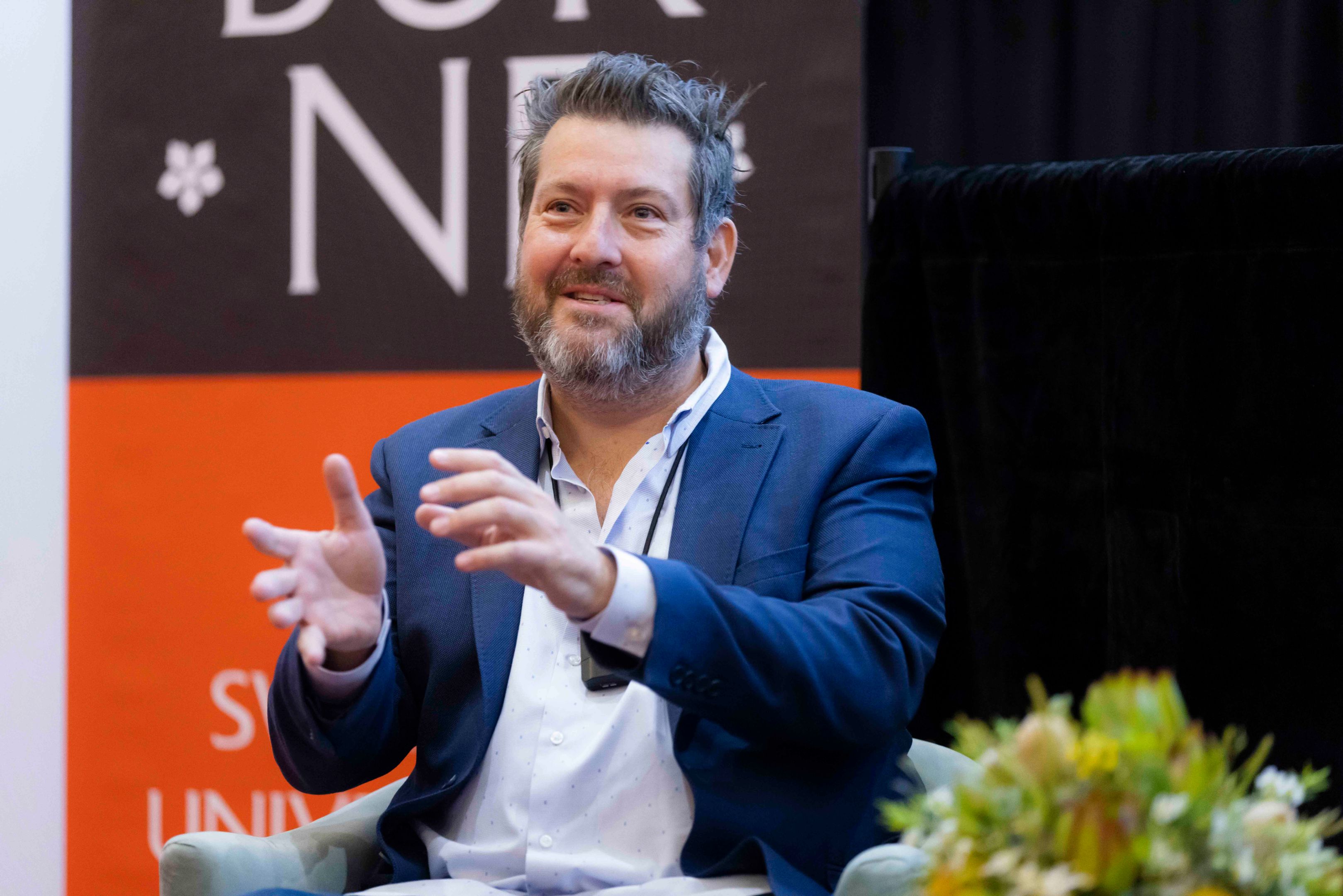 Man in blue blazer and white shirt sits in armchair in front of Swinburne banner as he delivers AGSE Oration