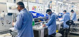 Scientists in blue lab coats, masks and screen protector shields work in a laboratory surrounded by machinery
