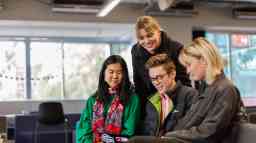 A group of young female and male students huddle together inside whilst looking at reading material.