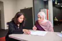 Two females sit at a desk as one helps the other through a book 