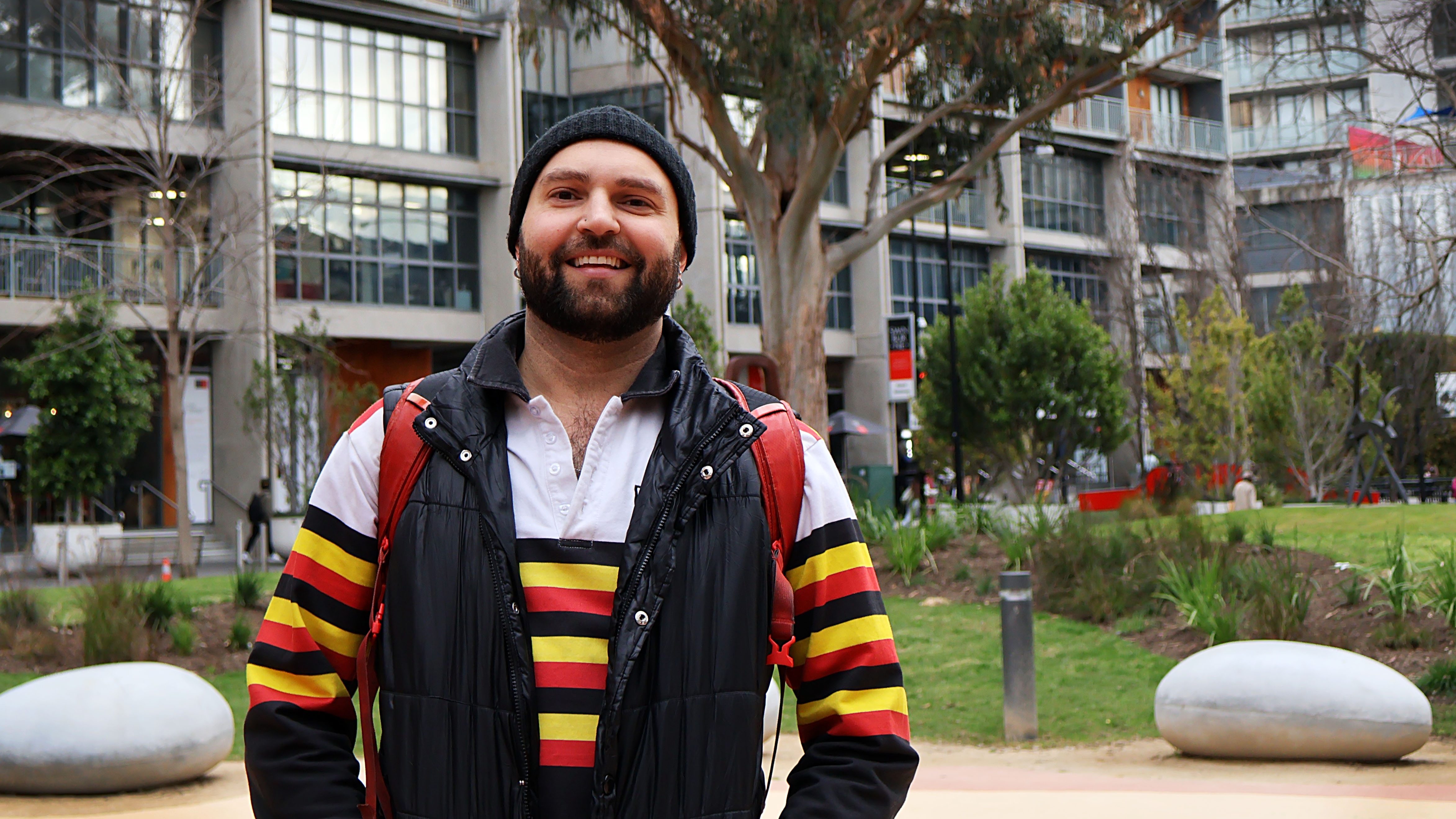 Tony Ryder is standing in the Indigenous Learning Circle. He is wearing a beaning and a shirt with black, yellow and red stripes. Behind him is some greenery and some of the campus buildings.