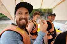 A young man holds a tropical fish while sitting on a boat wearing a life jacket. In the background are two other young men wearing life jackets and smiling at him. 