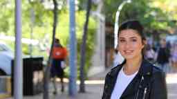 A young female with black hair tied in a pony tail, wearing a leather jacket and white tshirt, smiles at camera.