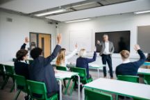 Rear view of secondary students in uniform sitting at desks, teacher standing in front teaching.