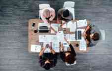 Team of 5 business people collaborating around a table with their laptops and paperwork.