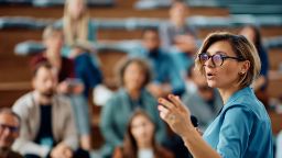 A female teacher wearing glasses is speaking in front of a lecture theatre of students.