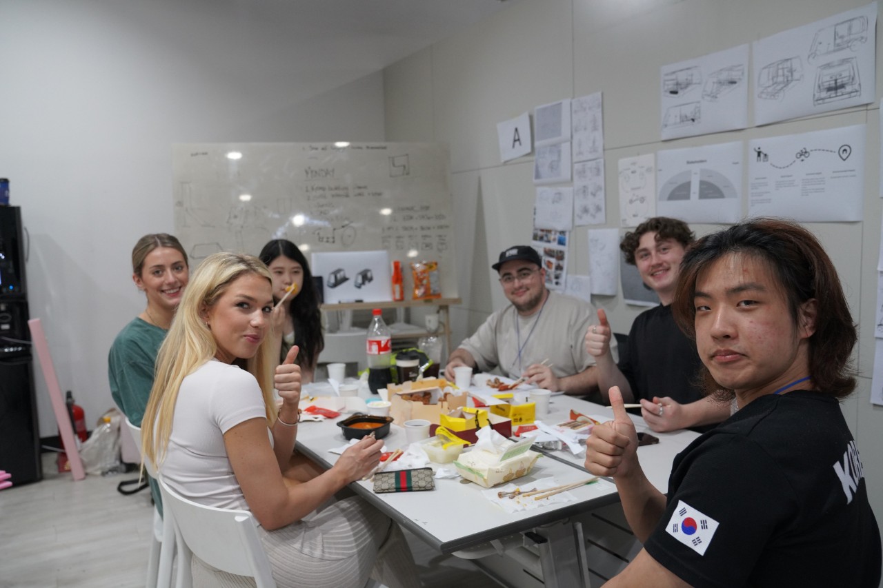 A group of students wit around a table eating takeway with design sketches on the walls behind them 